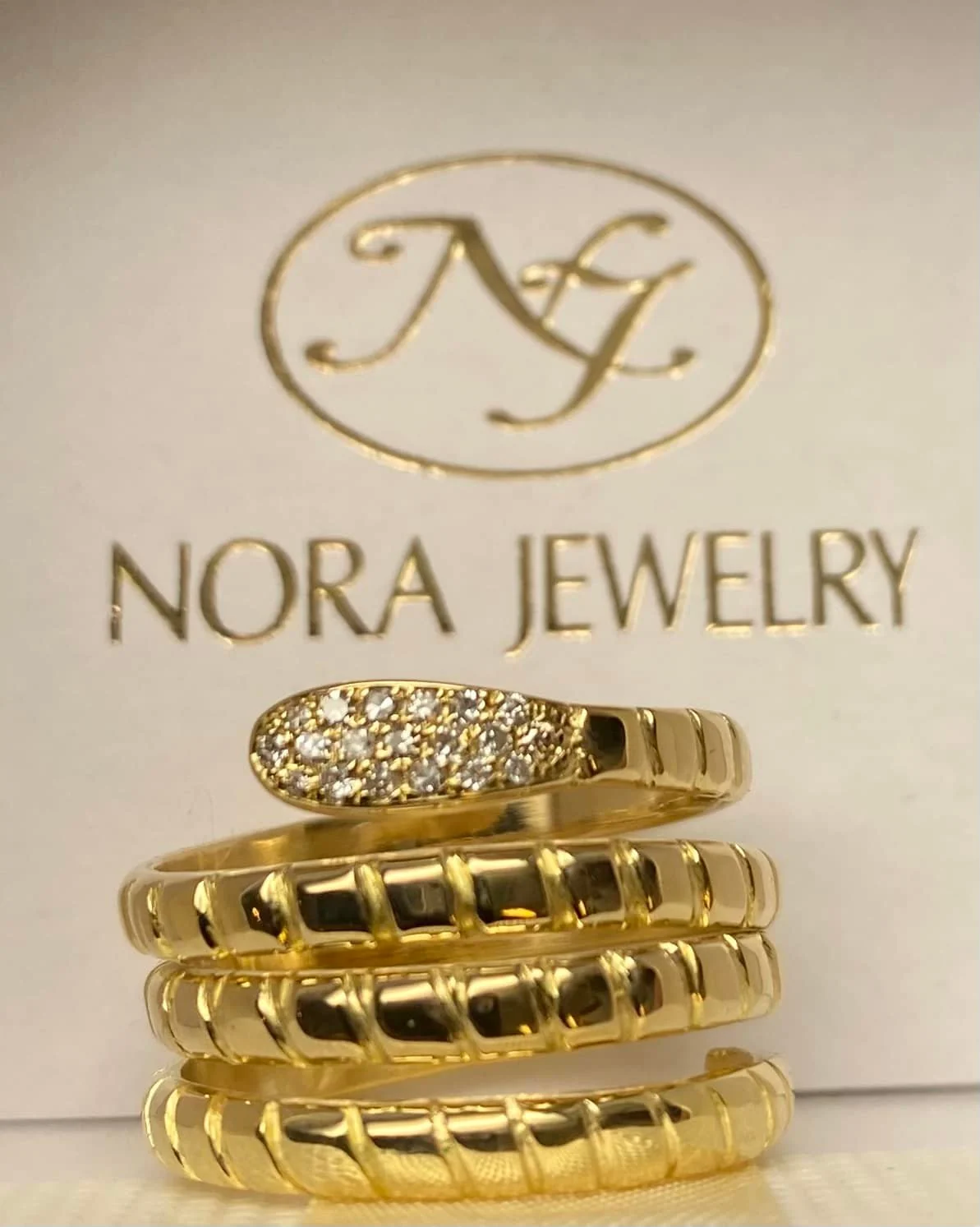 Anello in oro giallo a spirale con pavé di diamanti, davanti al marchio Nora Jewelry in rilievo dorato