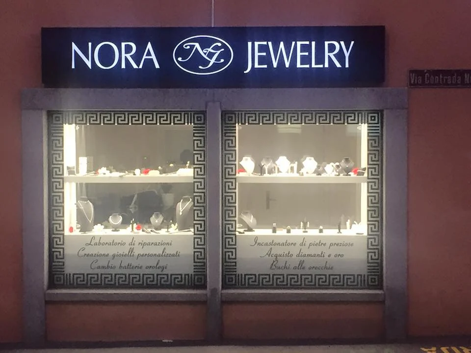 Le due vetrine illuminate di Nora Jewelry la sera, con l'insegna e la greca sui vetri